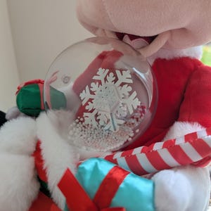 May include: A plush Mickey Mouse figure dressed in a Santa Claus outfit, holding a clear snow globe with a white snowflake design and white beads. The figure is wearing a red suit with white trim and holding a blue gift with a red bow.