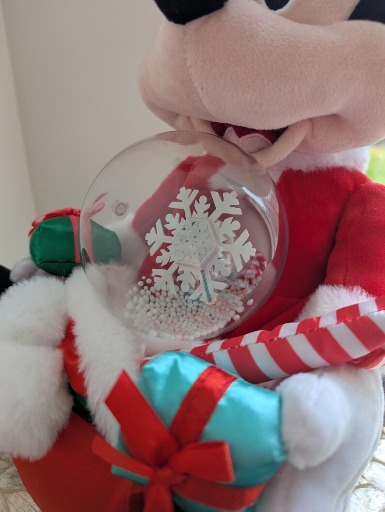 May include: A plush Mickey Mouse figure dressed in a Santa Claus outfit, holding a clear snow globe with a white snowflake design and white beads. The figure is wearing a red suit with white trim and holding a blue gift with a red bow.
