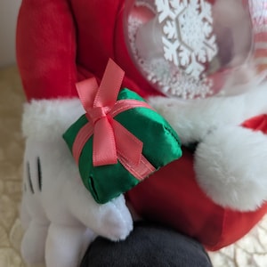 May include: A plush figure in a red Santa suit with white trim, holding a green gift box tied with a pink ribbon. A clear ornament with a white snowflake design is also visible. The figure's white gloved hand is holding the gift.