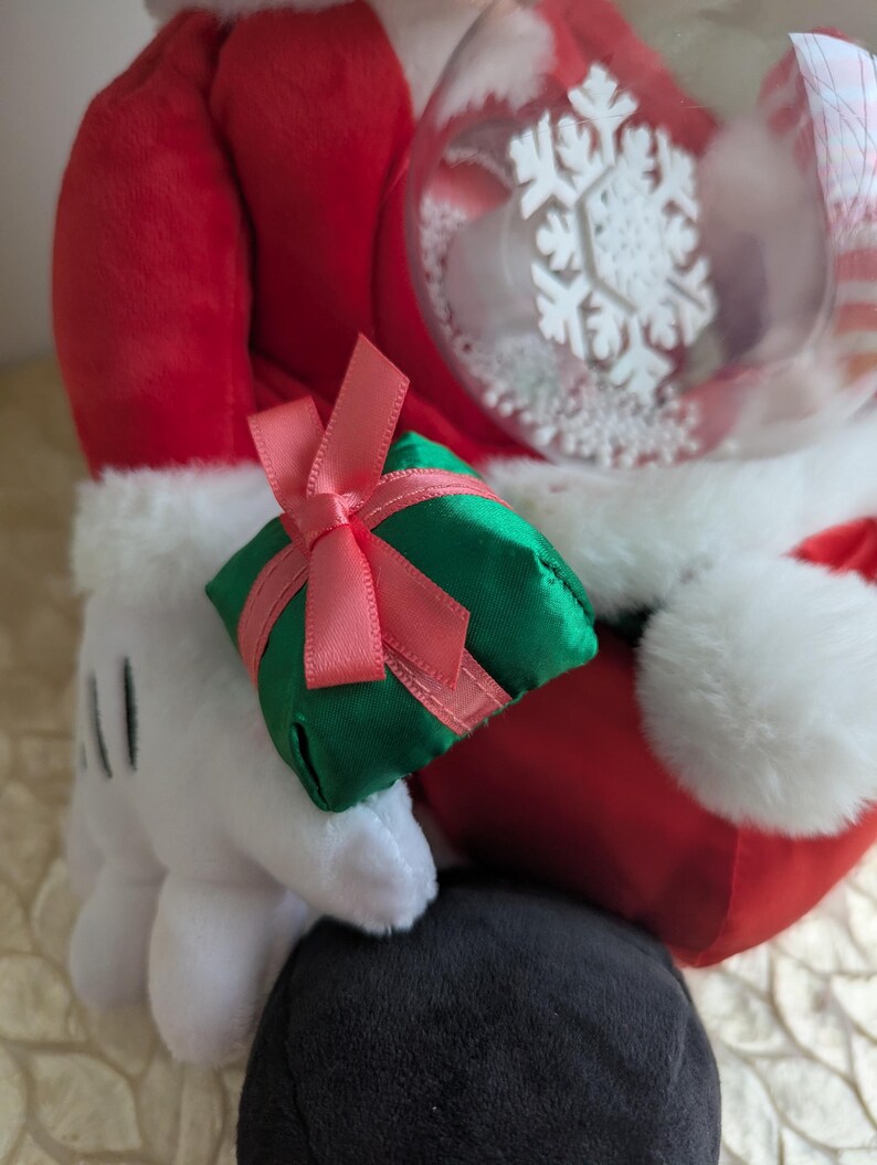 May include: A plush figure in a red Santa suit with white trim, holding a green gift box tied with a pink ribbon. A clear ornament with a white snowflake design is also visible. The figure's white gloved hand is holding the gift.