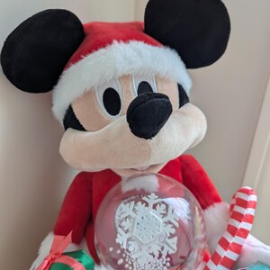 May include: A plush Mickey Mouse dressed in a Santa Claus outfit, complete with a red hat and coat trimmed with white faux fur. Mickey holds a snow globe with a snowflake design and a candy cane.