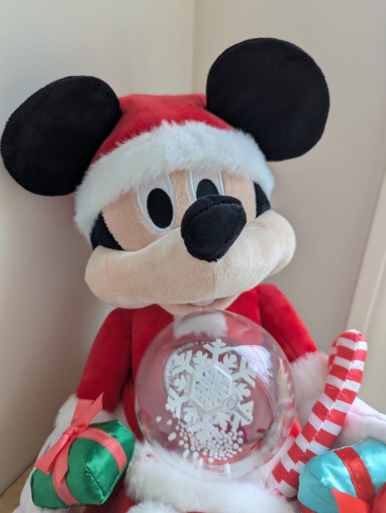 May include: A plush Mickey Mouse dressed in a Santa Claus outfit, complete with a red hat and coat trimmed with white faux fur. Mickey holds a snow globe with a snowflake design and a candy cane.