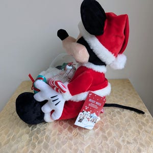 May include: A plush Mickey Mouse figure dressed in a Santa Claus outfit, holding a snow globe with candy canes. The figure is red, black, and white, with a Santa hat and gloves. A tag with the words "Mickey Mouse" is attached.