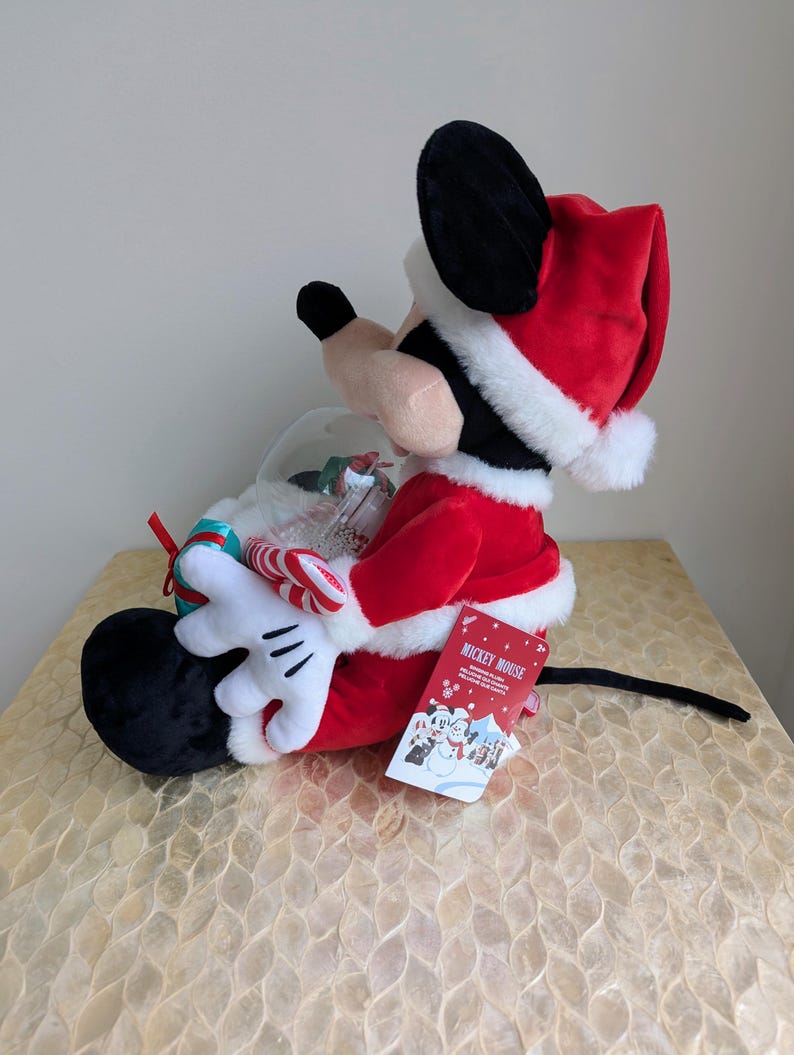 May include: A plush Mickey Mouse figure dressed in a Santa Claus outfit, holding a snow globe with candy canes. The figure is red, black, and white, with a Santa hat and gloves. A tag with the words "Mickey Mouse" is attached.