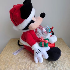May include: A plush Mickey Mouse dressed as Santa Claus, wearing a red suit and hat with white trim. Mickey holds a clear globe filled with candy canes and a small green gift box with a red bow. The plush toy has black ears and a white gloved hand.