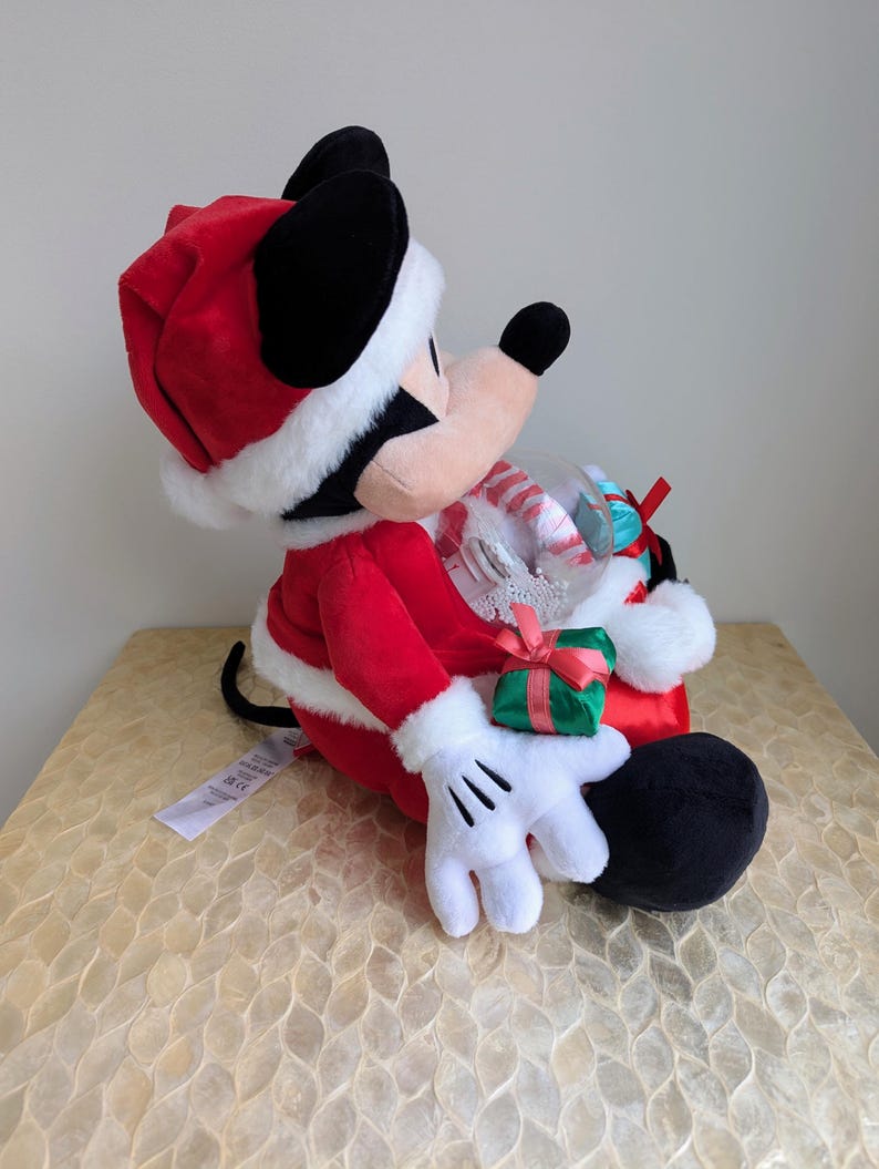May include: A plush Mickey Mouse dressed as Santa Claus, wearing a red suit and hat with white trim. Mickey holds a clear globe filled with candy canes and a small green gift box with a red bow. The plush toy has black ears and a white gloved hand.