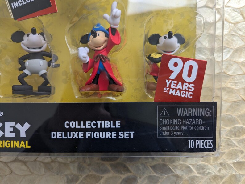 Puede incluir: Set de figuras de lujo coleccionables con tres figuras de Mickey Mouse. Una figura es en blanco y negro, otra est&aacute; vestida de hechicero y la tercera lleva un atuendo amarillo y rojo. El embalaje incluye una pegatina "90 Years of Magic".