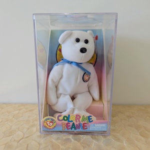 May include: A white plush bear with black eyes and a black nose, wearing a blue ribbon around its neck. The bear is inside a clear plastic display case labeled Color Me Beanie with a Ty logo. The bear features a small circular tag on its chest.