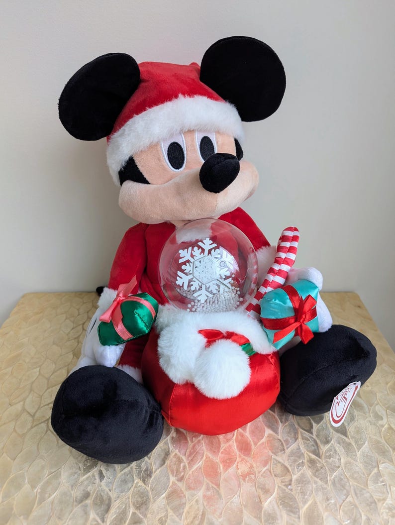 May include: A plush Mickey Mouse figure dressed in a Santa Claus outfit, complete with a red hat and suit with white trim. Mickey holds a snow globe with a snowflake design and small wrapped gifts.