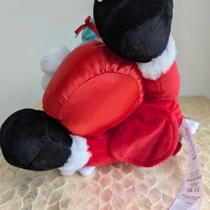May include: A plush Santa Claus doll, primarily red with black boots and a black hat. The doll has white faux fur trim and a small green gift bag. A tag is attached to the doll.