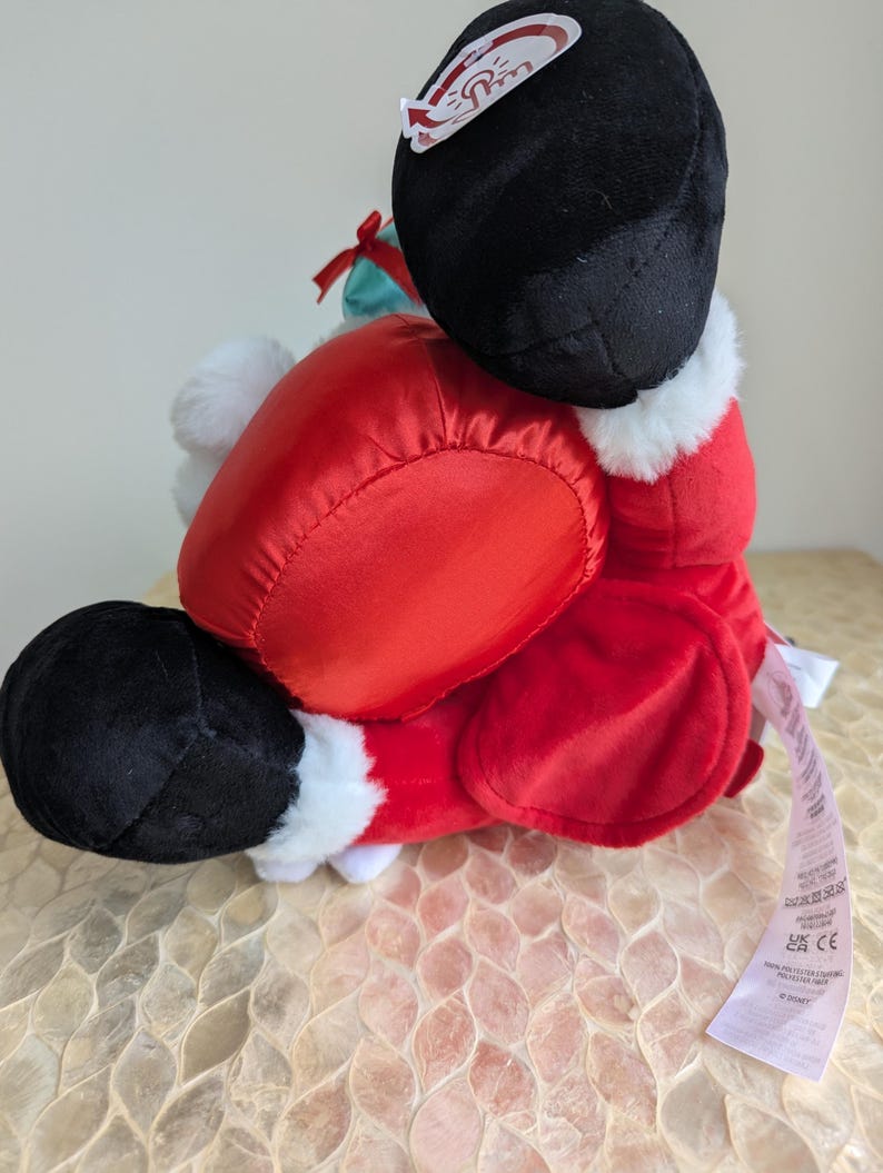 May include: A plush Santa Claus doll, primarily red with black boots and a black hat. The doll has white faux fur trim and a small green gift bag. A tag is attached to the doll.