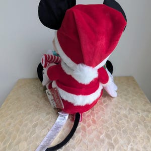 May include: A plush Mickey Mouse toy dressed in a Santa Claus outfit. The toy features a red Santa hat and suit with white trim, black ears, and a candy cane. A tag is attached to the toy.