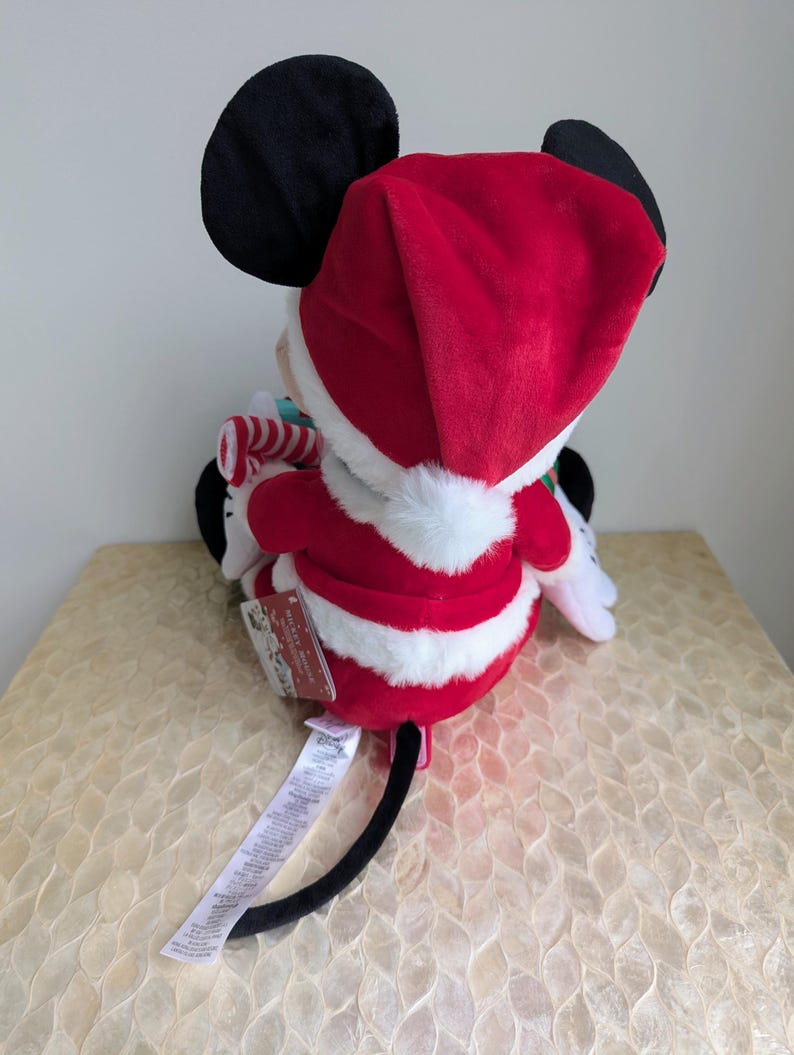 May include: A plush Mickey Mouse toy dressed in a Santa Claus outfit. The toy features a red Santa hat and suit with white trim, black ears, and a candy cane. A tag is attached to the toy.