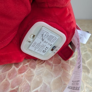 May include: A close-up of a plush red toy with a white battery compartment. The compartment has a switch and a label with text. A white tag with text is attached to the toy.