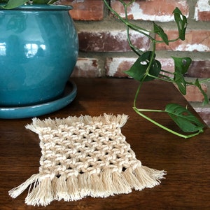 May include: A square, cream-colored macrame coaster with fringe on all sides. The coaster is sitting on a wooden surface.