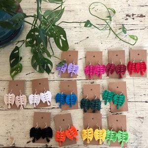 Puede incluir: Surtido de pendientes de macramé hechos a mano en varios colores, incluyendo rosa, morado, azul, verde, naranja y negro. Los pendientes se muestran en pequeñas tarjetas con fondo blanco.