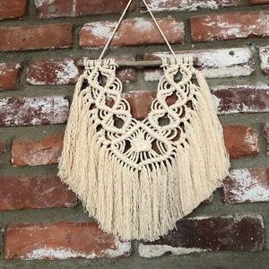 May include: A beige macrame wall hanging with a wooden dowel at the top and fringe at the bottom. The hanging is in the shape of a teardrop and has a geometric pattern.