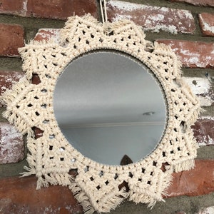 May include: A round mirror with a crocheted frame in a cream color. The frame has a fringe detail and is hanging on a brick wall.
