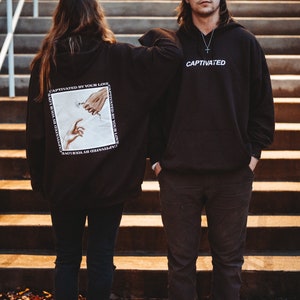 May include: Two people wearing black hoodies with white text. The hoodie on the left has a graphic of two hands reaching towards each other with a flower in between. The text on the hoodie says "CAPTIVATED BY YOUR LOVE". The hoodie on the right says "CAPTIVATED".