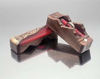 Op de afbeelding: Twee houten ringendoosjes, waarvan één open, met een ring op een rode en geruite stof. De doosjes zijn gemaakt van donker hout met een unieke, natuurlijke nerf. Eén doosje heeft een rode harsinleg.