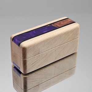 Könnte beinhalten: Eine rechteckige Holzkiste mit hellem Holzfinish. Die Oberseite hat einen Streifen aus tiefviolettem Harz und einen Abschnitt aus braunem Holz. Die Kiste ist geschlossen, mit einer sichtbaren Naht. Die Kiste spiegelt sich in der darunter liegenden Oberfläche.