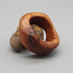 May include: A wooden ring with a natural wood grain finish. The ring is shaped like a circle with a gap in the middle. The ring is sitting on top of a smaller, round, brown stone with white spots.