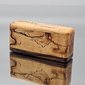 Pode incluir: Uma caixa retangular de madeira com uma parte superior castanha clara e uma base castanha escura. A parte superior apresenta um padrão único de linhas castanhas escuras, que se assemelham a uma cordilheira. A caixa é refletida na superfície abaixo.