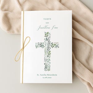 Könnte beinhalten: Ein weißes Büchlein mit dem Wort "TAUFE" oben und dem Namen "Jonathan Finn" in eleganter Schrift. Ein Kreuz aus grünen Blättern ist darunter zentriert. Der Text "St. Aurelia Meisenheim" und ein Datum stehen unten. Eine beige Schnur bindet das Büchlein.