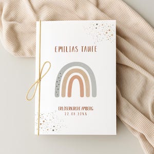 May include: A white card with the text "EMILIAS TAUFE" above a rainbow illustration in neutral tones. The card is tied with a gold string and features additional text and decorative speckles.