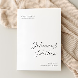 Könnte beinhalten: Eine weiße Hochzeitseinladung mit schwarzer Schrift. Der Text lautet oben "Willkommen zur Trauung von", in der Mitte die Namen "Johanna & Sebastian" und unten das Datum und der Ort.