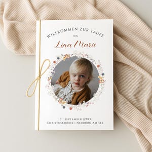 Könnte beinhalten: Eine weiße Einladungskarte mit dem Text "WILLKOMMEN ZUR TAUFE" und "Lina Marie" in einer dekorativen Schriftart. Ein Foto eines Kindes ist von einem Blumenkranz umgeben. Die Karte ist mit einer goldenen Schnur gebunden.