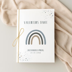 Könnte beinhalten: Eine weiße Karte mit dem Text "VALENTINS TAUFE" über einer Illustration eines blauen, grauen und braunen Regenbogens. Die Karte ist mit einer goldenen Schnur gebunden. Der Text "ERLÖSERKIRCHE AMBERG 28.08.20XX" ist darunter gedruckt.