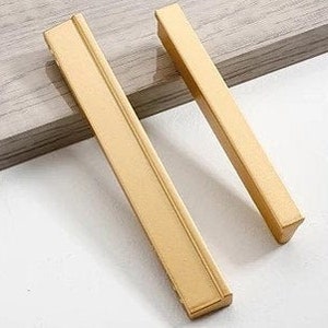 May include: Two gold-toned cabinet pulls with a rectangular shape and a slightly angled edge. The pulls are made of metal and have a brushed finish.