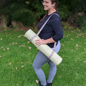 May include: A person wearing grey leggings and black shoes is carrying a light green yoga mat with a brown and white patterned strap.