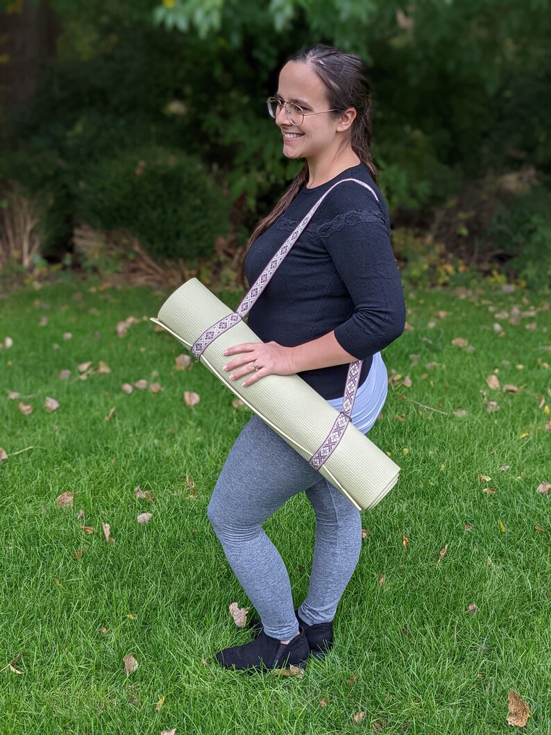 May include: A person wearing grey leggings and black shoes is carrying a light green yoga mat with a brown and white patterned strap.