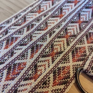 May include: A white woven belt with a geometric pattern in shades of brown, pink, and orange. The belt has a metal ring on one end.