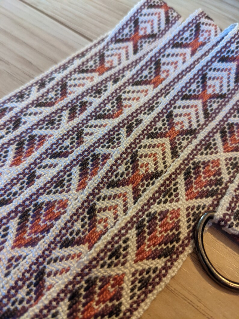 May include: A white woven belt with a geometric pattern in shades of brown, pink, and orange. The belt has a metal ring on one end.