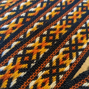 May include: Close-up of a woven fabric with a geometric pattern in black, orange, white, and yellow.