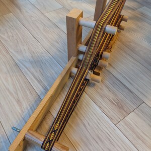 May include: A wooden loom with a colorful woven band in shades of yellow, orange, black, and white. The loom is made of natural wood and has a simple design.