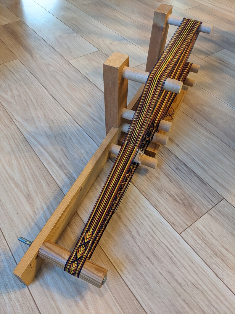 May include: A wooden loom with a colorful woven band in shades of yellow, orange, black, and white. The loom is made of natural wood and has a simple design.