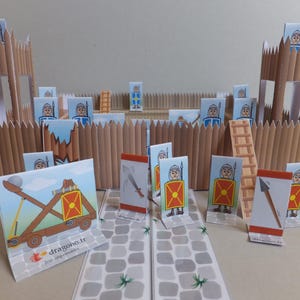 Peut inclure: Un fort en carton avec une clôture en bois et des tours. Il y a plusieurs petites figurines en papier de soldats avec des boucliers et des épées. Il y a une catapulte et une échelle devant le fort. Le texte "dragono.fr" est imprimé sur la catapulte.