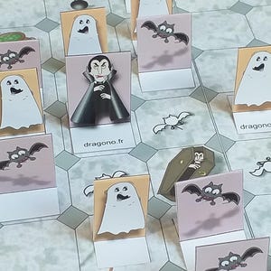 Peut inclure: Un plateau de jeu avec différentes cartes sur le thème d'Halloween. Les cartes présentent des images de fantômes, de chauves-souris, d'un vampire et d'un cercueil. Les cartes sont sur un fond gris clair avec un motif hexagonal. Le texte "dragono.fr" apparaît sur deux des cartes.