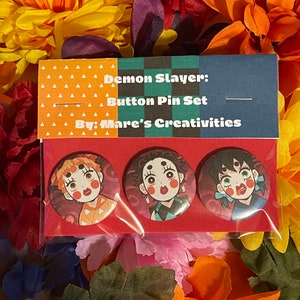 May include: Three Demon Slayer button pins featuring anime characters with red, green, and yellow accents. The pins are displayed on a red background with a white label that reads "Demon Slayer: Button Pin Set By: Mare's Creativities".