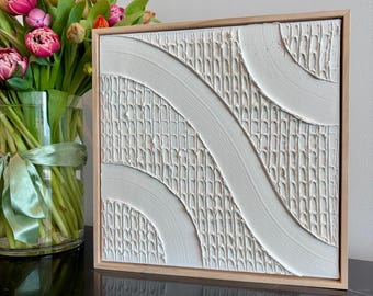 Op de afbeelding: Een vierkant wandkunstwerk met een wit gestructureerd oppervlak, verhoogde diagonale lijnen en een rasterachtig patroon, gevat in een lichtgekleurde houten lijst. Het kunstwerk toont een combinatie van gladde rondingen en verticale ribbels.