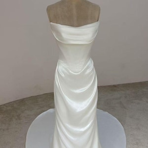 Può includere: Un abito da sposa in raso bianco su un manichino. Il corpetto senza spalline ha uno scollo piegato e una vita aderente. La gonna sfocia in un lungo strascico. Il manichino ha il numero 2.