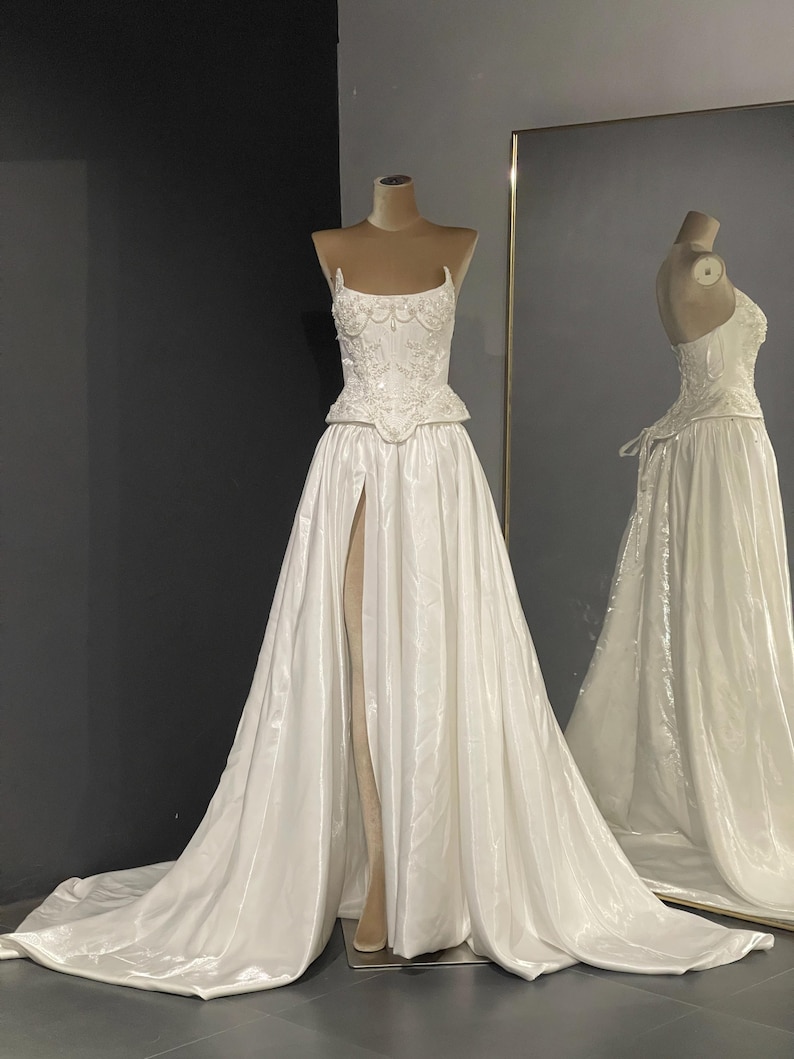 May include: A white wedding dress on a mannequin. The dress features a fitted bodice with intricate beading and a high slit skirt. The dress is displayed in front of a mirror, reflecting the back of the gown.