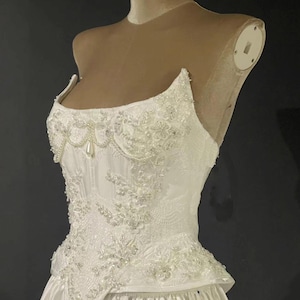May include: White wedding dress with a fitted bodice and a full skirt. The bodice is embellished with intricate floral lace, beads, and pearls. The dress is displayed on a mannequin.