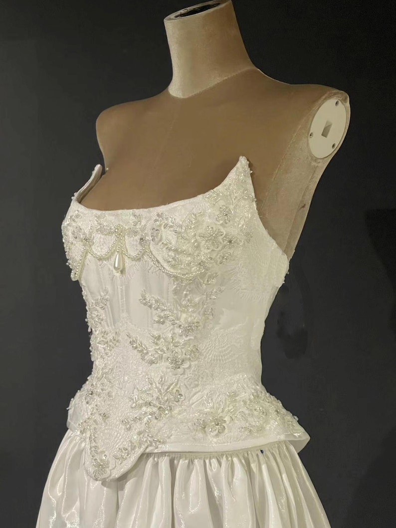 May include: White wedding dress with a fitted bodice and a full skirt. The bodice is embellished with intricate floral lace, beads, and pearls. The dress is displayed on a mannequin.