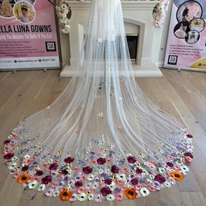 May include: A long, sheer white bridal veil with a floral embroidered border. The veil is displayed on a mannequin and features a detailed design of colorful flowers in shades of red, orange, pink, and purple.