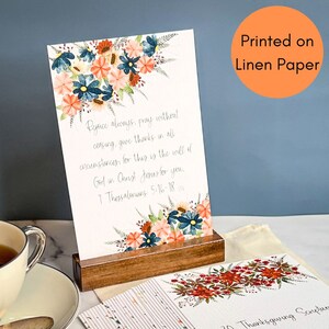 May include: A set of 20 floral-themed cards with a scripture quote printed on each card. The quote reads "Rejoice always, pray without ceasing, give thanks in all circumstances, for this is the will of God in Christ Jesus for you. 1 Thessalonians 5:16-18". The cards are printed on linen paper and come with a wooden stand.